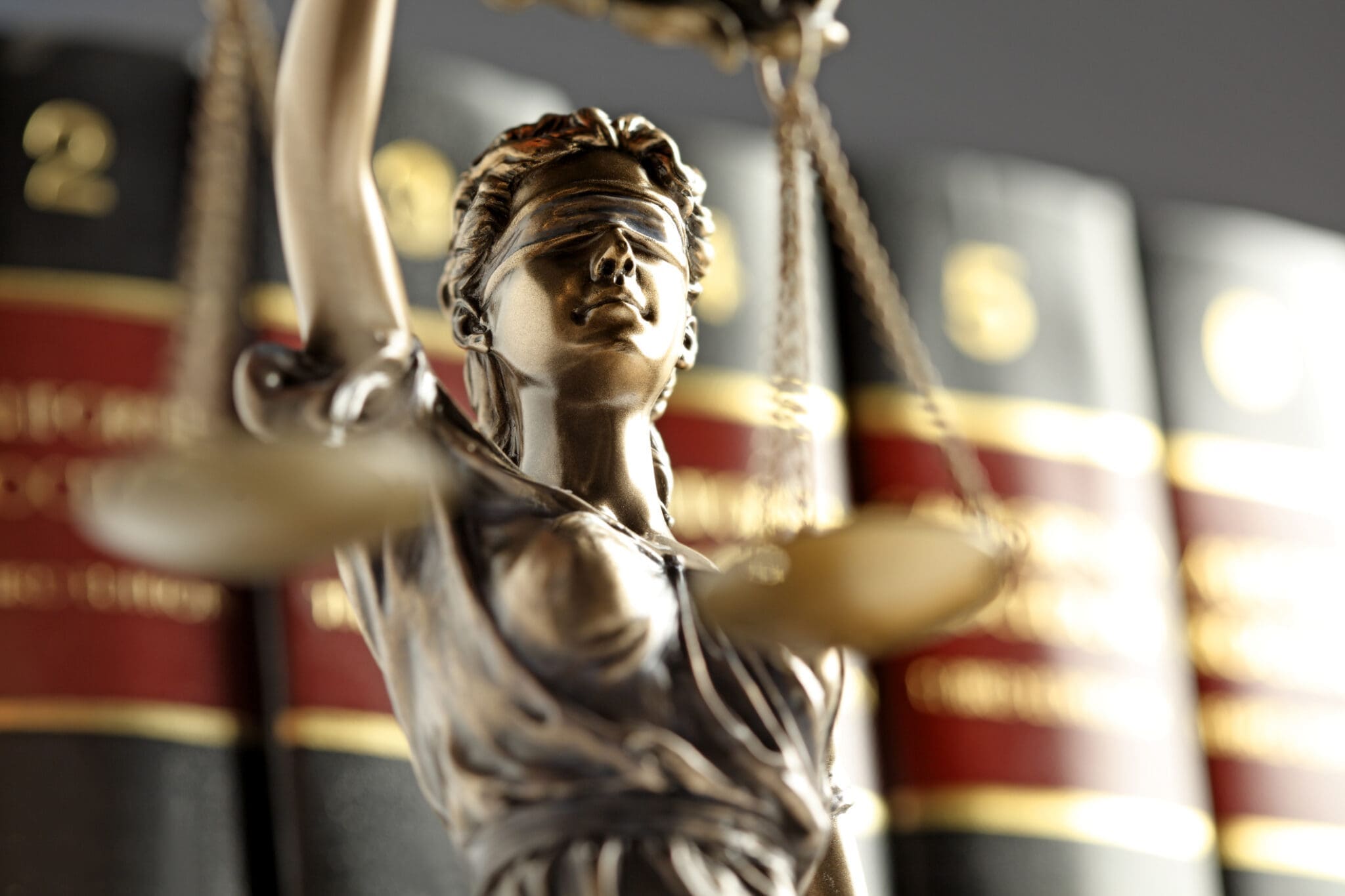 A close up of the statue of lady justice standing in front of an out of focus row of law books. The law books are bound in red and black leather with gold writing.